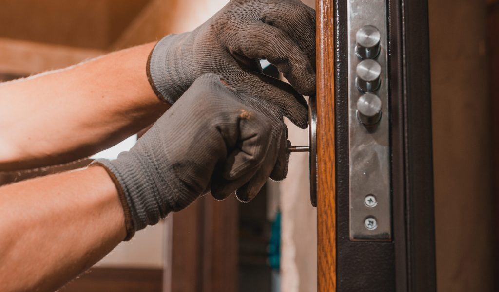 Close-up, the carpenter installs a custom lock in the front metal door, using a drill and hammer and other tools.2020